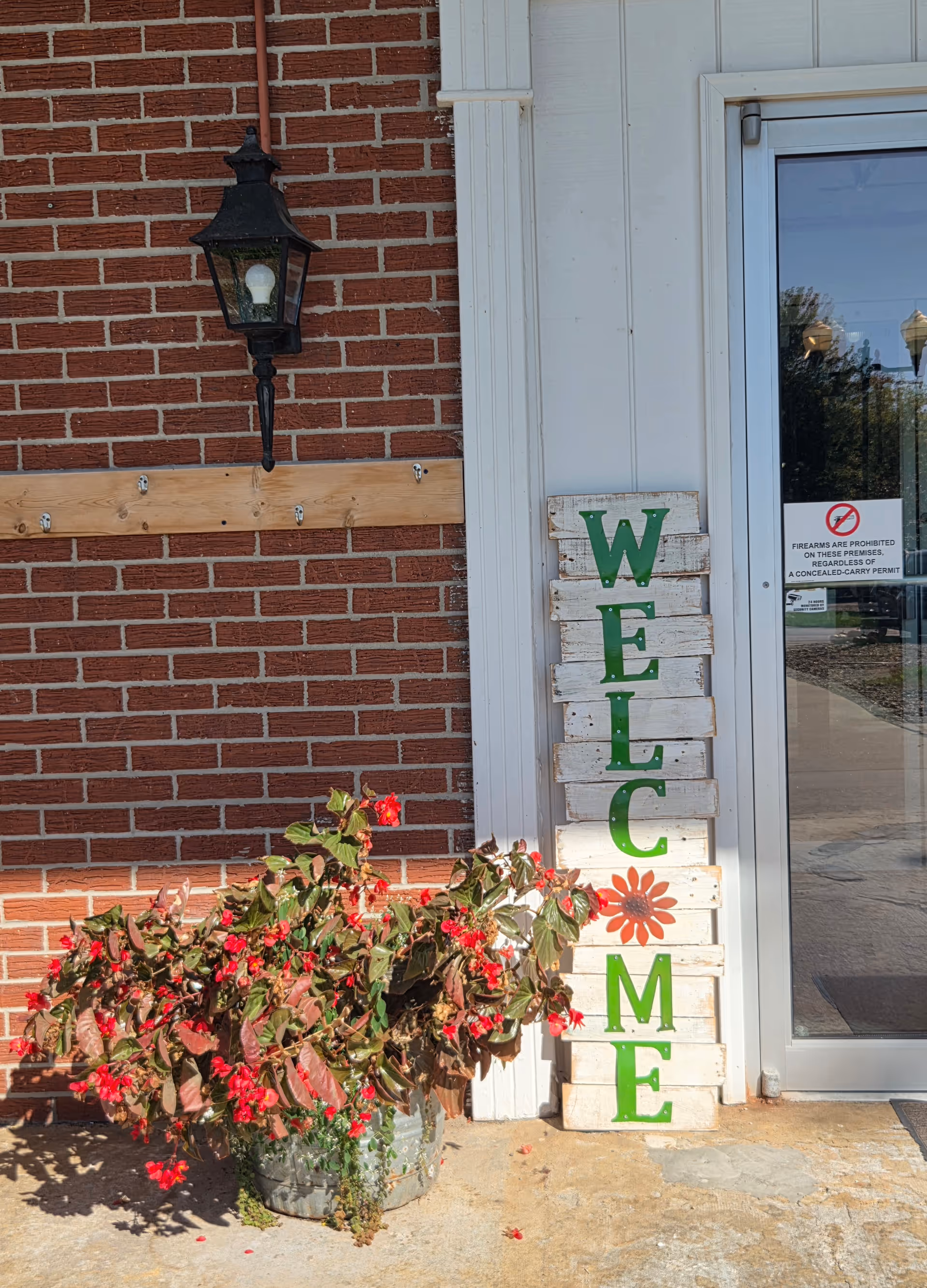 Entrance area showing a vertical wooden "WELCOME" sign leaning by a glass door, a potted plant with red flowers, a brick wall, and a wall-mounted lantern.