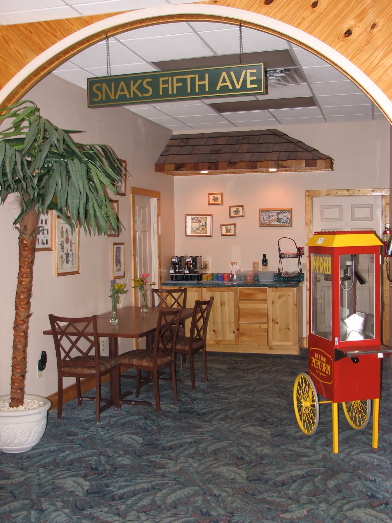 Interior view of a small dining and snack area with a wooden table and four chairs, a potted artificial palm tree, a popcorn machine on yellow wheels, and a counter with coffee machines and cups. A sign hanging from the ceiling reads 'SNAKS FIFTH AVE'. The walls have framed pictures and the floor is carpeted with a leaf pattern.