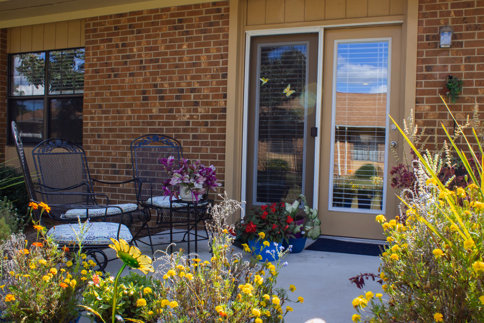 Brick-front patio entrance with a glass door, metal chairs and cushions, and colorful potted flowers.