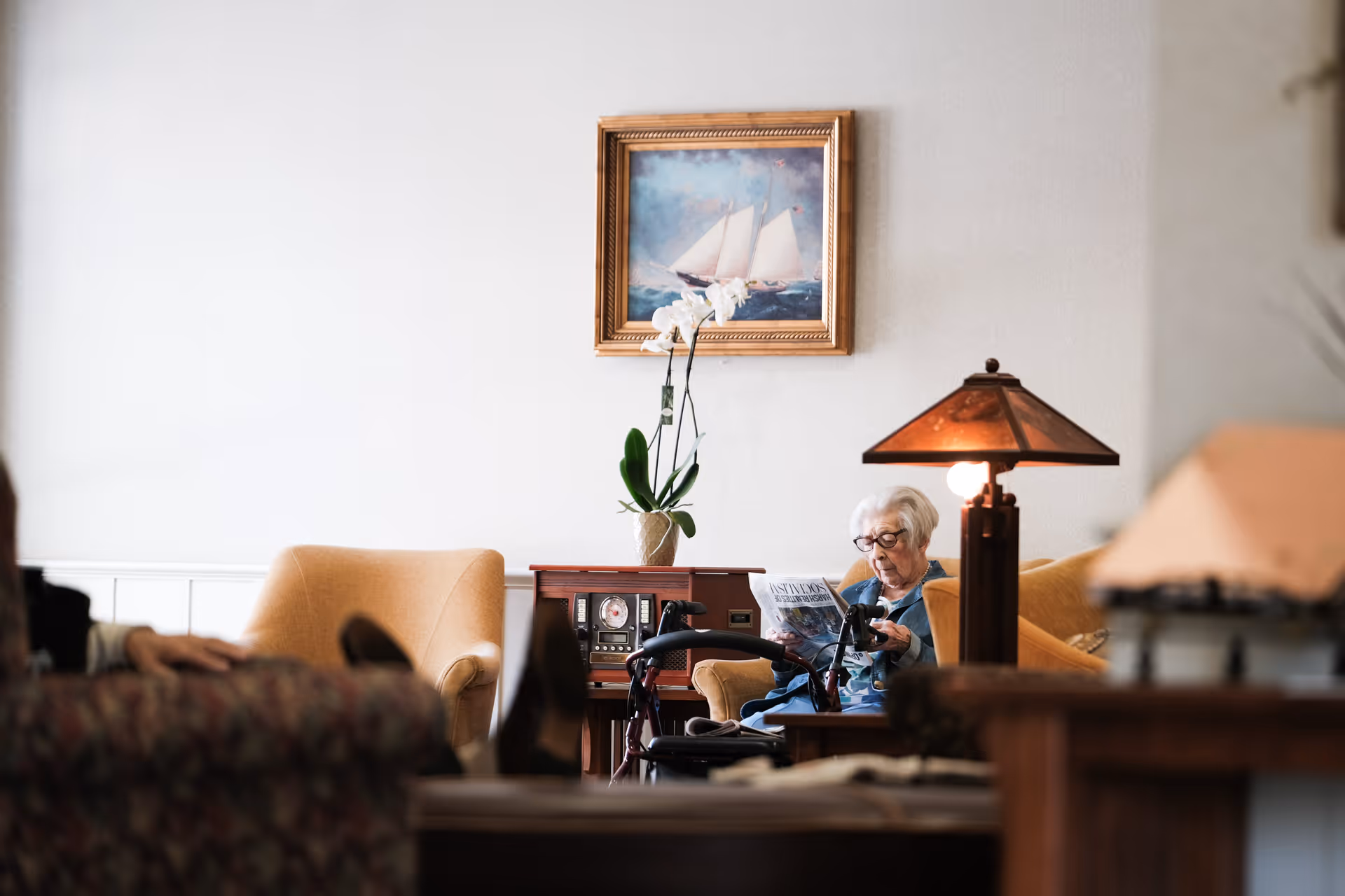 An elderly woman sitting in a cozy living room area reading a newspaper. The room features comfortable armchairs, a wooden side table with a lamp, a vintage radio, a potted orchid, and a framed painting of a sailboat on the wall.