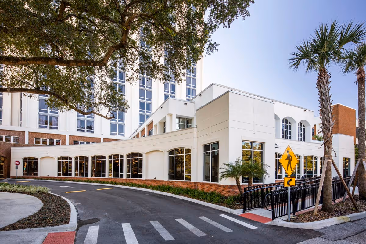 Exterior view of Westminster Towers, showing a curved driveway with a pedestrian crosswalk, a white multi-story building with large windows, palm trees, and a stop sign under a clear blue sky.