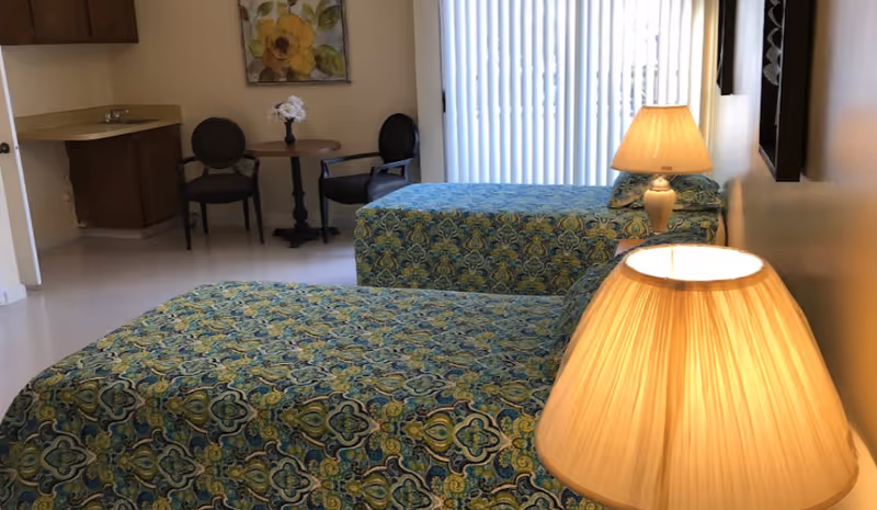 Two twin beds with green patterned covers, bedside lamps, a small table with chairs, and a sink area in an assisted living bedroom with vertical blinds.