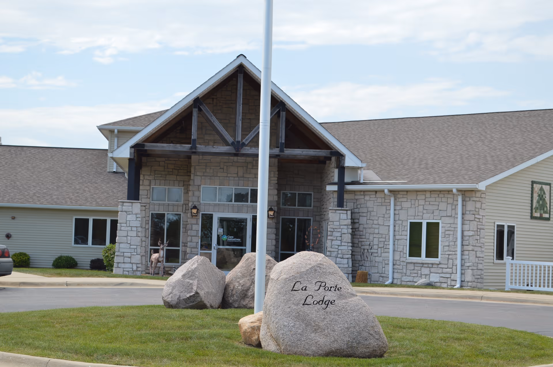 Front exterior view of a building with stone and siding facade, a peaked roof entrance supported by wooden beams, and a large rock in the foreground engraved with 'La Porte Lodge'. A flagpole stands in front of the rock, and there is a driveway and some greenery around the building.