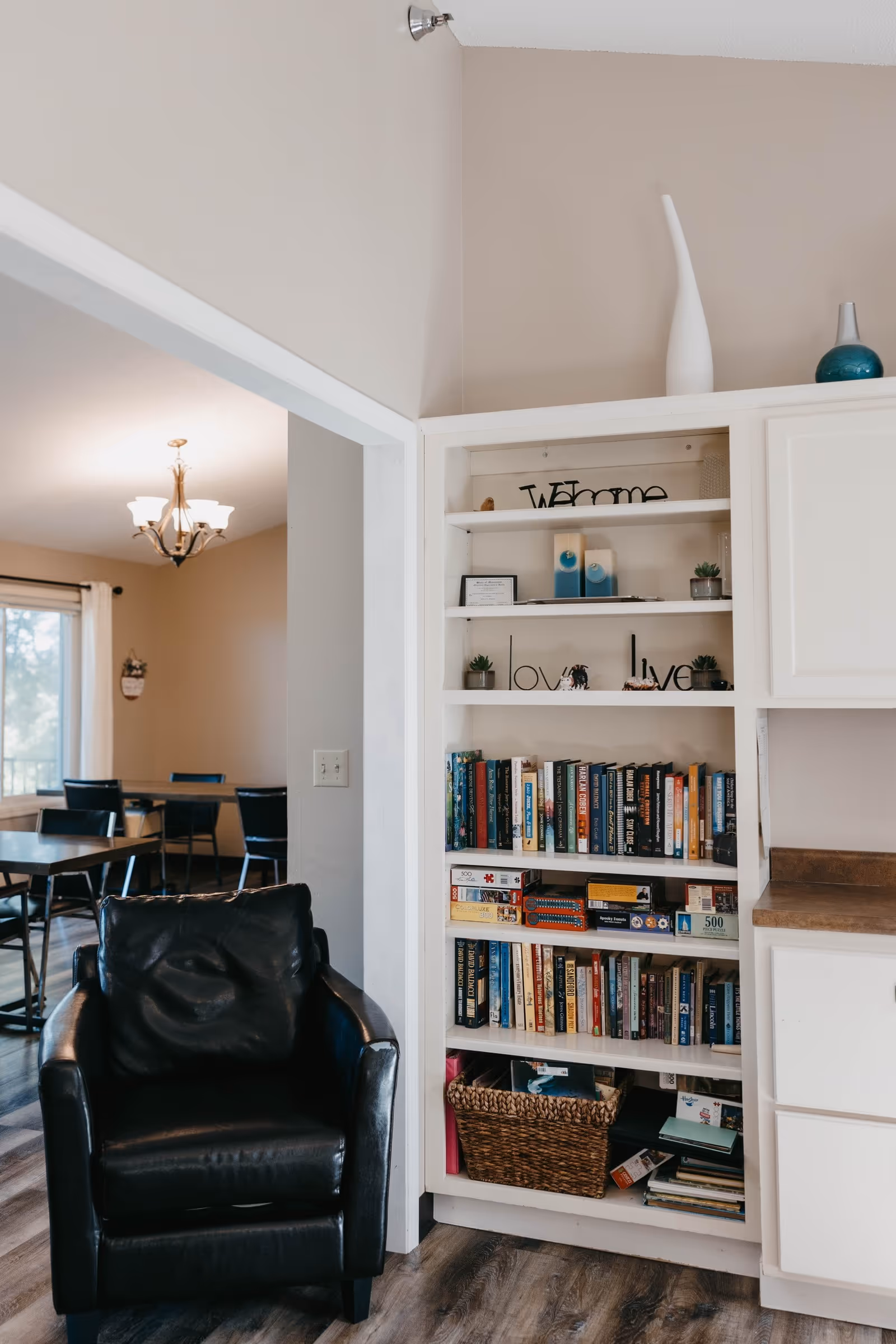 A cozy interior showing a black leather armchair beside built-in white bookshelves filled with books and decorative items, with a dining area visible in the background.