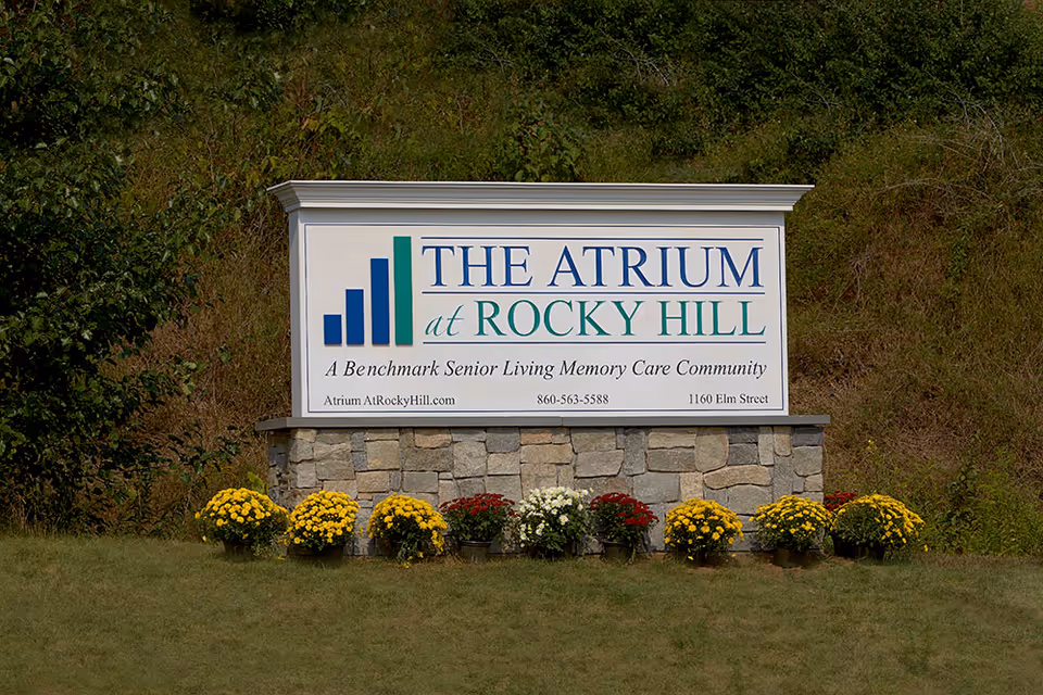 A stone-base roadside sign reading 'The Atrium at Rocky Hill, A Benchmark Senior Living Memory Care Community' with potted flowers in front and a grassy hill behind.