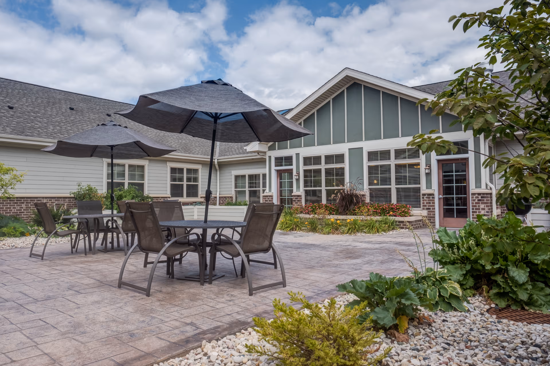 Patio courtyard with round tables, chairs and large umbrellas in front of a single-story memory care building and landscaped beds.