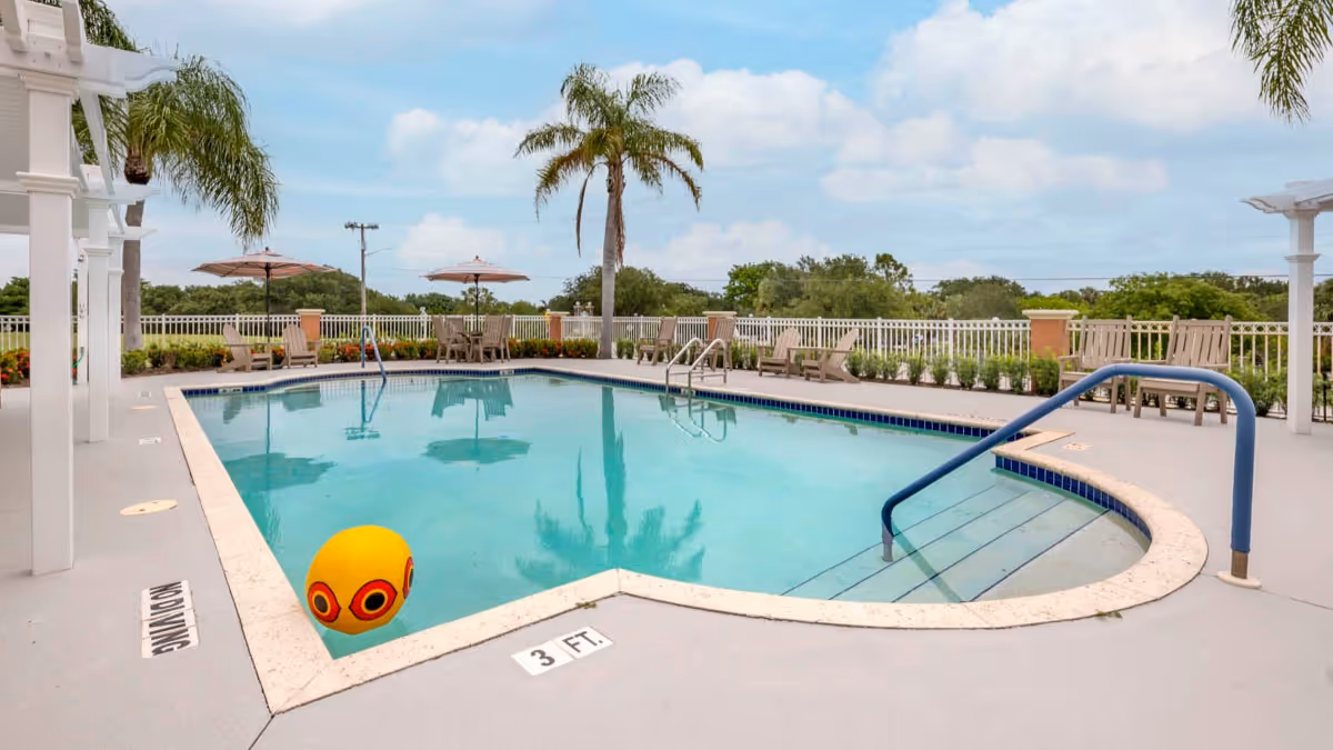 Outdoor swimming pool with a yellow and red beach ball floating in the water. The pool area is surrounded by a white fence, palm trees, lounge chairs, and umbrellas under a partly cloudy sky.