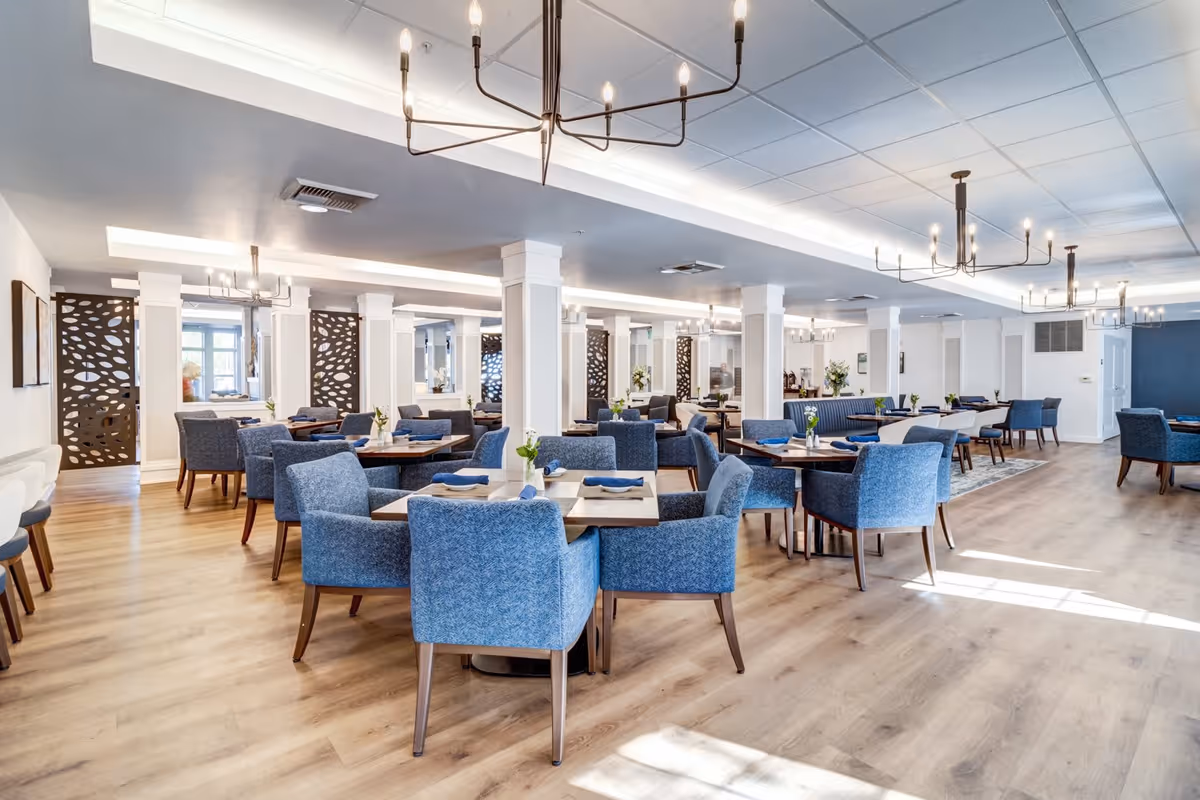 A spacious and well-lit dining room with multiple tables surrounded by blue upholstered chairs. The room features wooden flooring, modern chandeliers, decorative wooden panels, and small flower arrangements on each table.