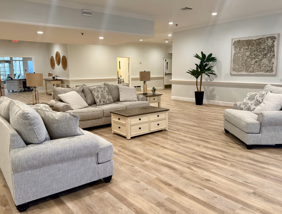A spacious and well-lit living room area with light wood flooring, featuring two gray sofas and one gray armchair arranged around a wooden coffee table with drawers. The room has neutral-colored walls, a potted plant, two table lamps on side tables, and decorative wall art. In the background, there is a hallway and additional furniture visible.