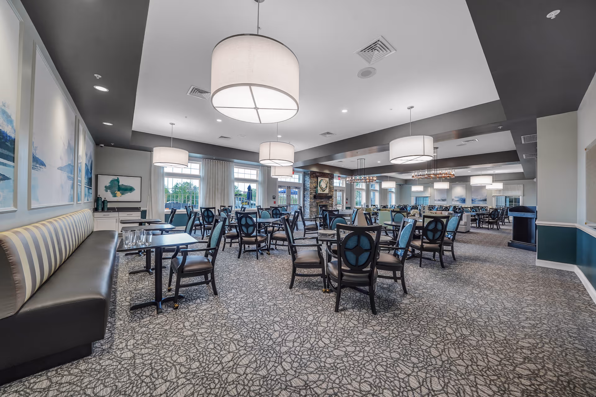 A spacious and well-lit dining room with multiple tables and chairs arranged neatly. The room features large windows allowing natural light, modern hanging light fixtures, patterned carpet, and artwork on the walls. There is a long cushioned bench along one side and a stone fireplace in the background.