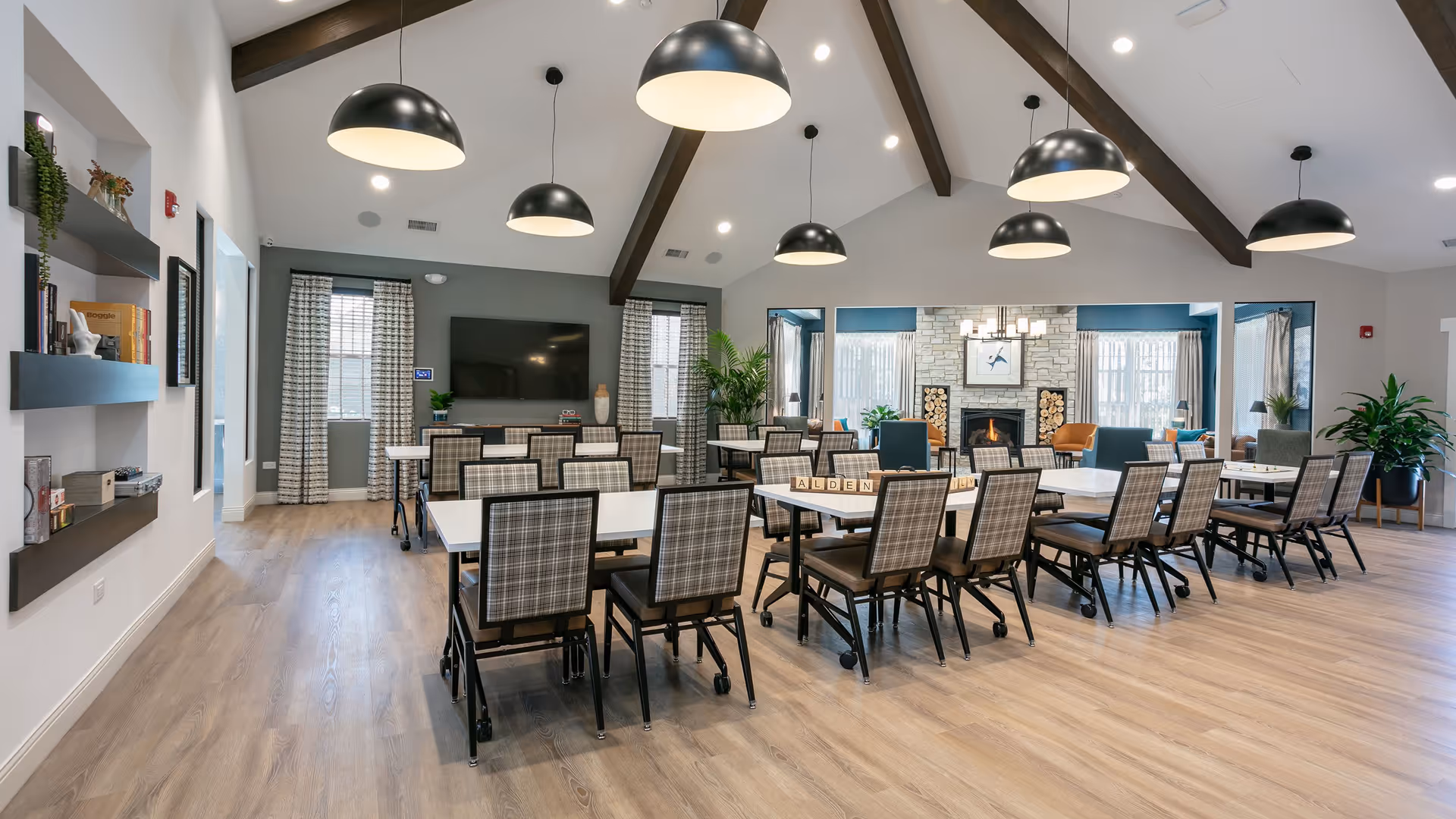 A spacious and well-lit dining room with multiple tables and plaid upholstered chairs arranged neatly. The room features a high ceiling with exposed wooden beams and large black pendant lights hanging down. At the far end, there is a cozy fireplace with stacked firewood on either side, flanked by windows with curtains. The floor is light wood, and there are plants and shelves with decorative items along the walls.