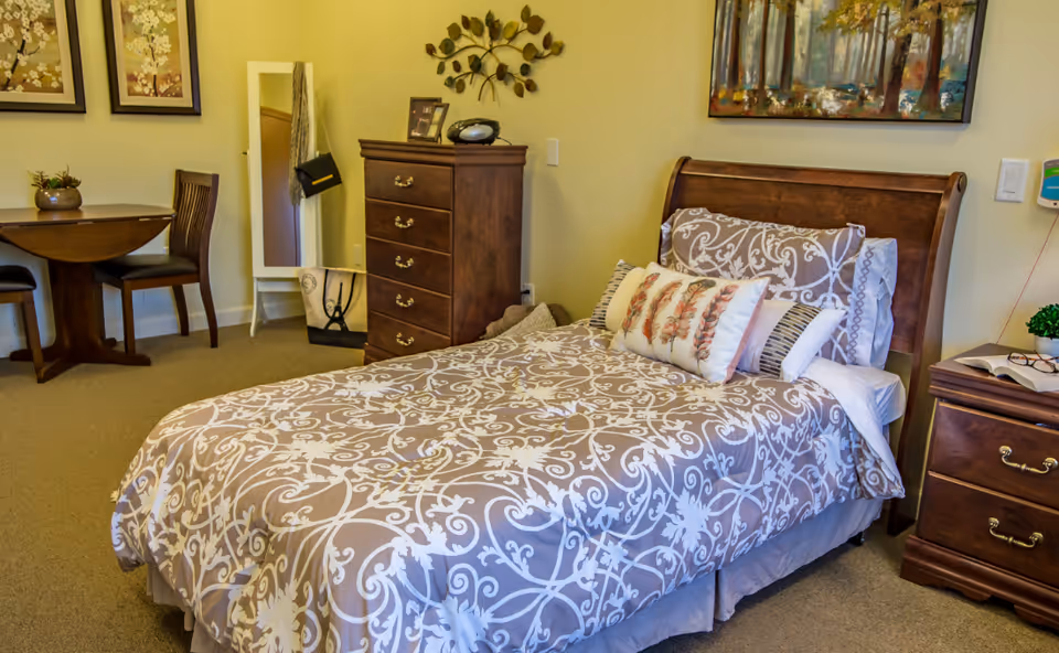 A cozy bedroom in a memory care community featuring a single bed with patterned bedding and multiple pillows. The room includes wooden furniture such as a chest of drawers, a nightstand with a book and glasses, and a small round table with two chairs. There is a full-length mirror with a handbag hanging on it, decorative wall art, and a small plant on the table.
