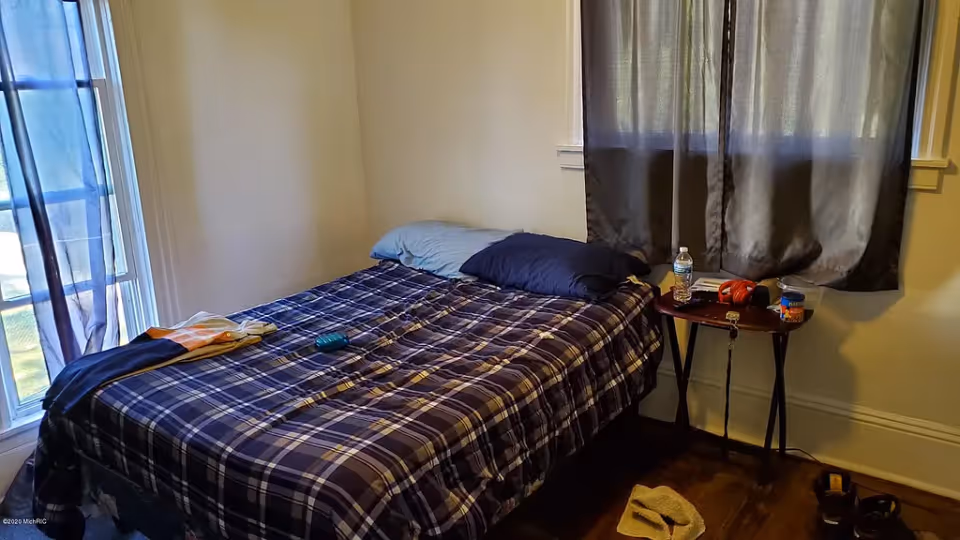 A small bedroom with a bed covered in a blue and white plaid blanket, two pillows, and a few personal items on the bed and a small table next to it. The room has two windows with dark curtains and wooden flooring.