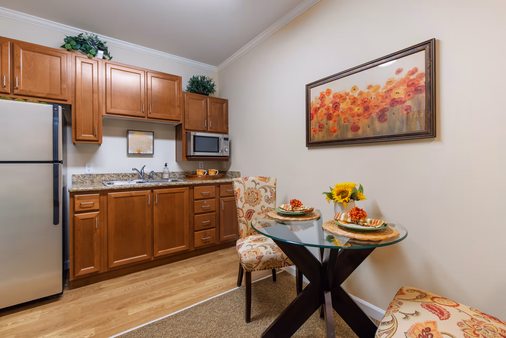 A small kitchen area with wooden cabinets, a stainless steel refrigerator, a microwave, and a granite countertop with a sink. Next to the kitchen is a glass-top dining table set for two with floral-patterned chairs. A framed painting of orange and red flowers hangs on the wall above the table.