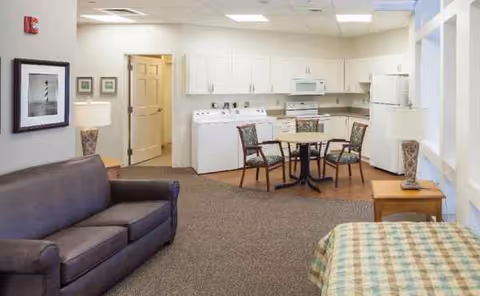 A senior living common area with a sofa, round table, and kitchen appliances in the background.
