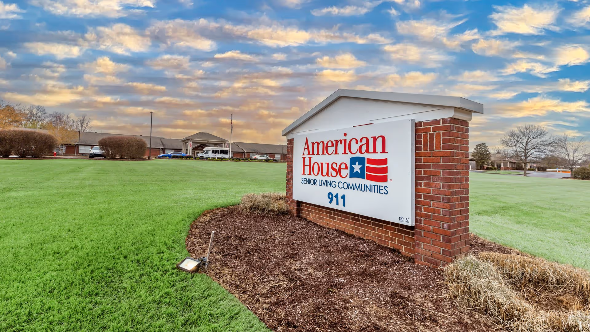 A large brick and white sign for American House Senior Living Communities with the number 911, set on a landscaped area with mulch and grass. In the background, there is a single-story building under a partly cloudy sky during sunset or sunrise.