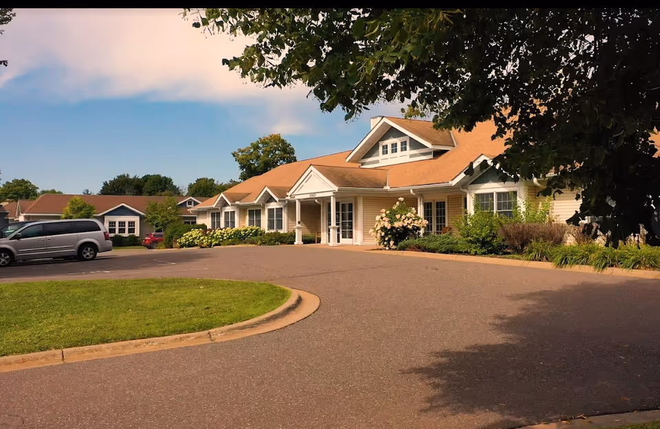 Single-story senior living building with a covered entrance, landscaped beds, a circular driveway and parked cars under a partly cloudy sky.