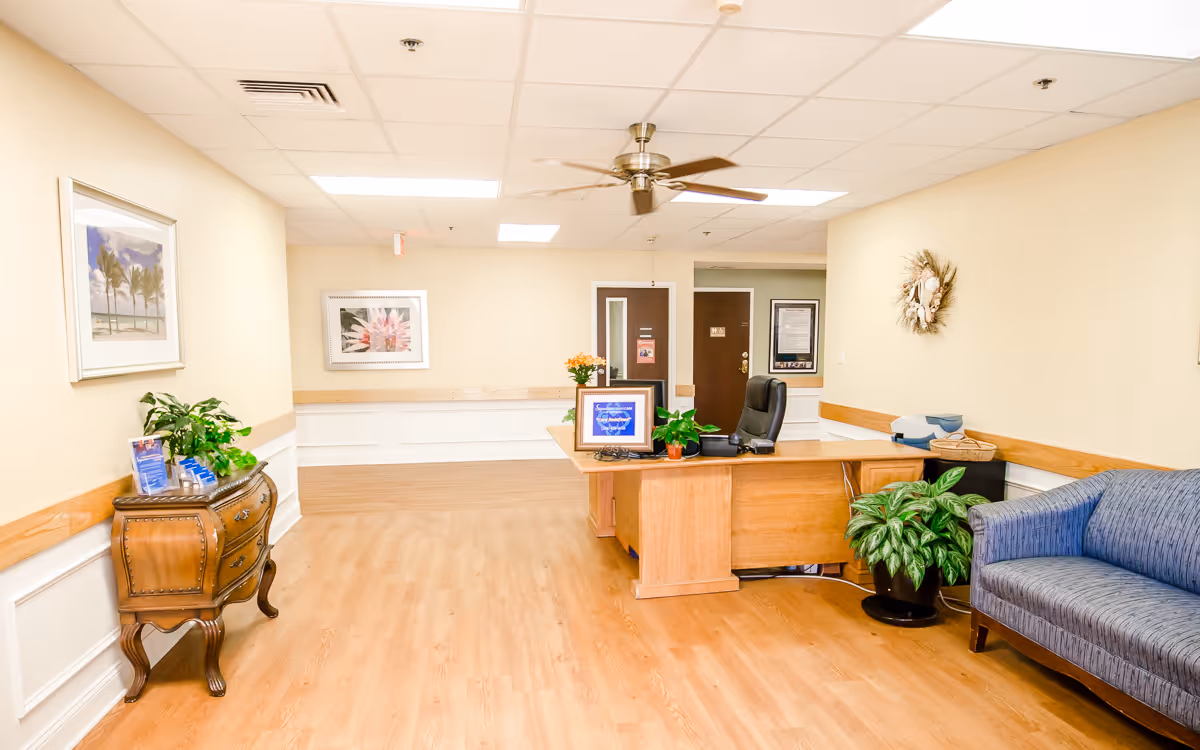 Reception area of a nursing facility with a wooden desk, seating, plants, artwork, and a ceiling fan.