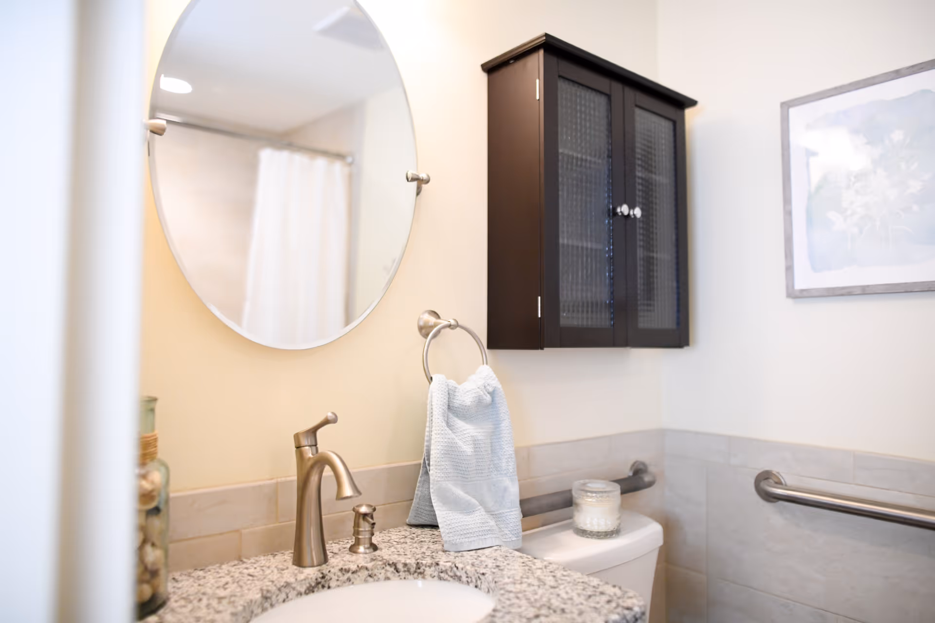 Accessible bathroom vanity with a round mirror, faucet, towel, wall cabinet and grab bars beside a toilet.