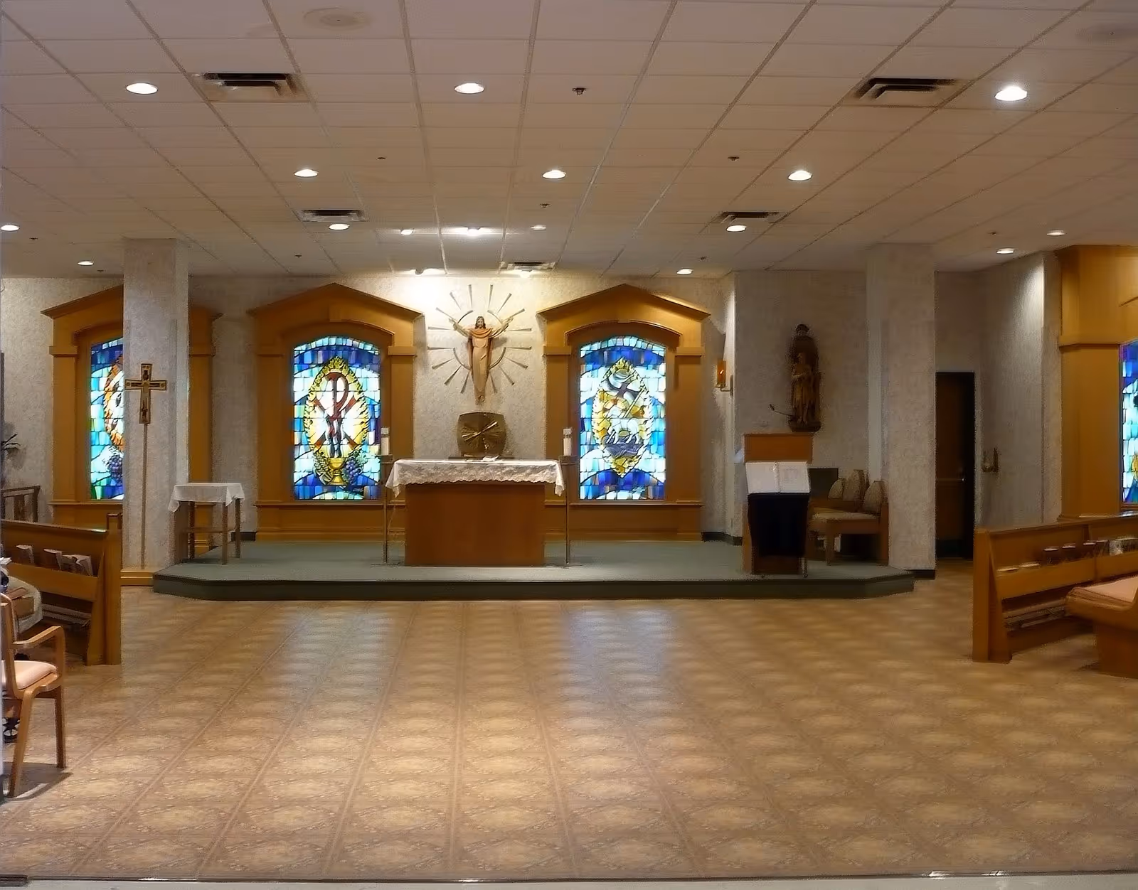 Interior view of a chapel with an altar in the center, flanked by stained glass windows and religious statues. Wooden pews are arranged on either side of the aisle, and the floor has a patterned tile design.