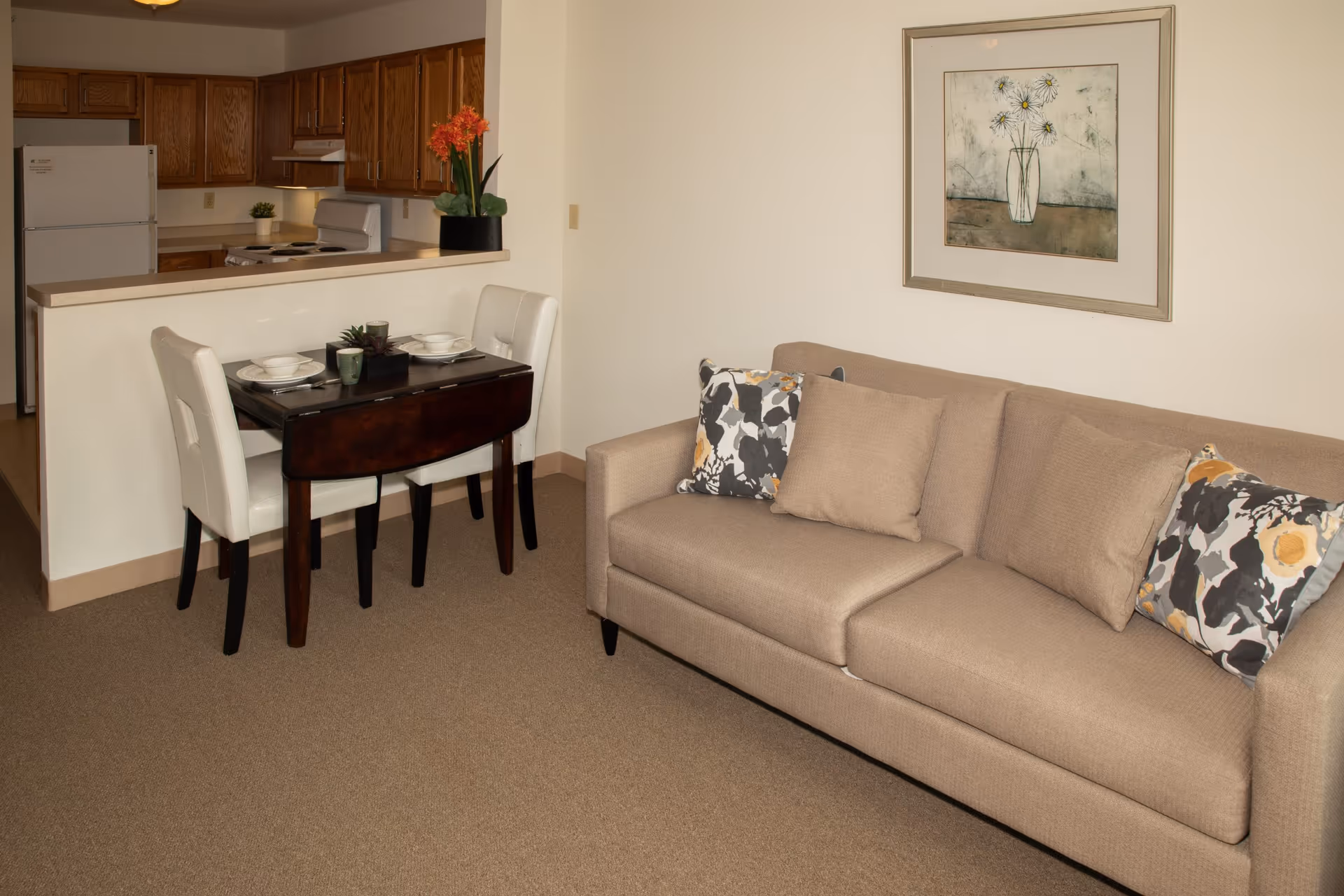 Small senior apartment living area with a beige sofa, patterned pillows, a two-seat dining table, and a kitchenette in the background.