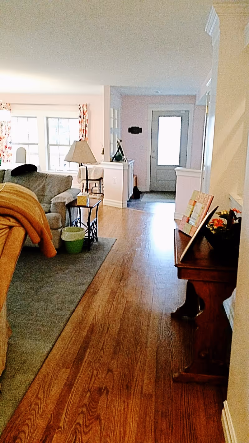 Interior view of a senior living facility showing a hallway with wooden flooring leading to a door with a window. On the left side, there is a living area with a gray couch, a side table with a lamp, and a green waste bin. On the right side, there is a wooden console table with framed pictures and a flower arrangement. The walls are light-colored, and there are windows with floral curtains in the background.