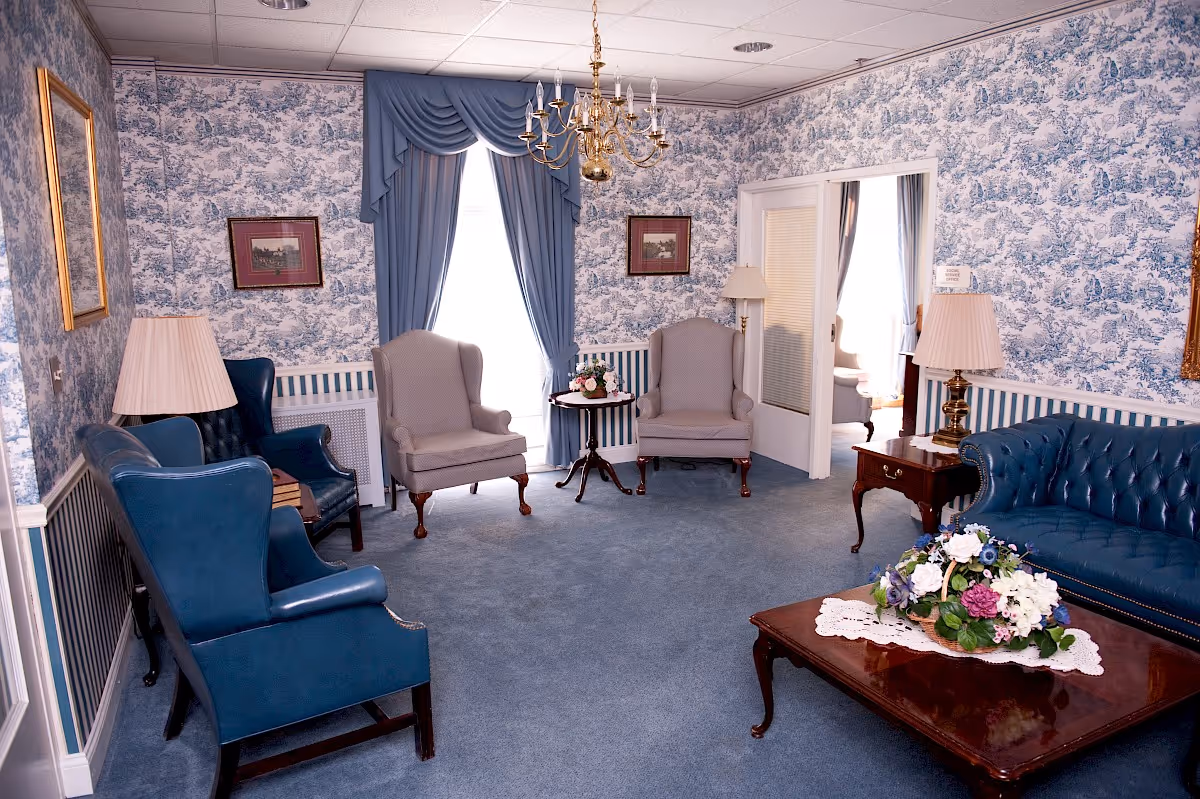 A traditional living room with blue and white toile wallpaper and matching blue carpet. The room features a blue leather sofa, two blue leather armchairs, two beige wingback chairs, a wooden coffee table with a floral centerpiece, side tables with lamps, and a chandelier hanging from the ceiling. Blue drapes frame a window in the background.