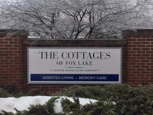 A brick sign for The Cottages of Fox Lake, a charter senior living community offering assisted living and memory care, with snow-covered bushes and a tree with frost in the background.