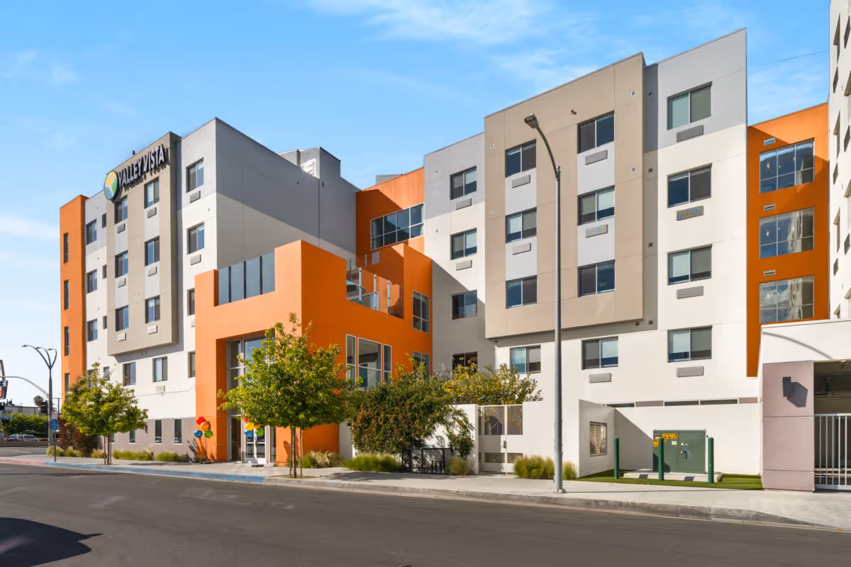 Street-level view of the modern multi-story Valley Vista Senior Living building with orange and white facades and small trees along the sidewalk.