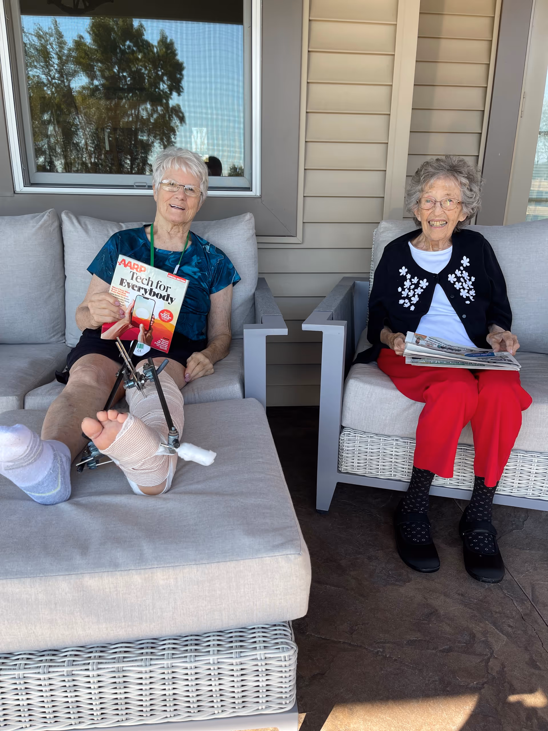 Two elderly women smiling while seated on a covered outdoor patio, one with a leg in a medical brace holding a magazine and the other holding a newspaper.