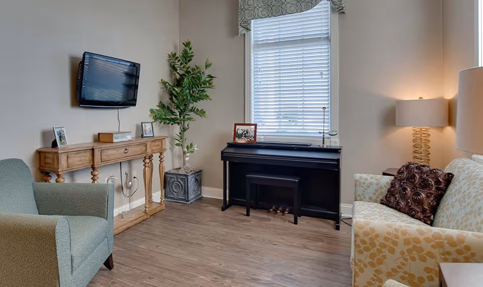 A cozy living room with a mounted flat-screen TV above a wooden console table displaying framed photos. There is a green armchair on the left, a potted plant in the corner, a black piano with a bench in front of a window with blinds, and a patterned sofa with a decorative pillow on the right. A table lamp with a spiral base is on a side table next to the sofa.