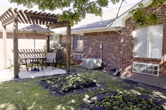 Outdoor patio area with a wooden pergola providing shade over a table and two chairs. The patio is adjacent to a brick building with windows and air conditioning units. There are small garden beds with plants and grass surrounding the patio, and tree branches providing additional shade.