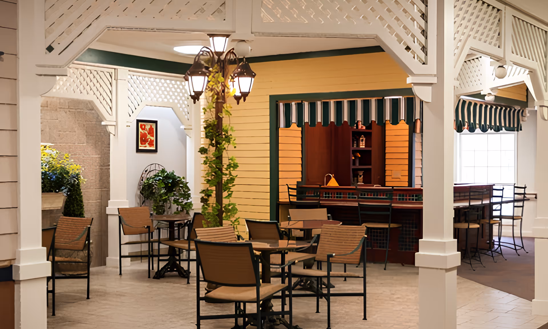 Indoor seating area with several tables and chairs arranged under a white lattice structure. A lamp post with three lanterns wrapped in green vines stands in the middle. In the background, there is a yellow wall with a green and white striped awning above a counter or bar area with stools. Potted plants and framed artwork decorate the space.