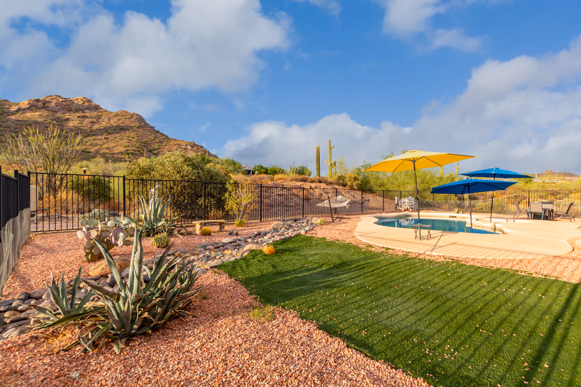 Outdoor area with a small swimming pool surrounded by a concrete deck, several colorful umbrellas providing shade, a hammock, desert landscaping with cacti and rocks, and a mountain in the background under a partly cloudy sky.