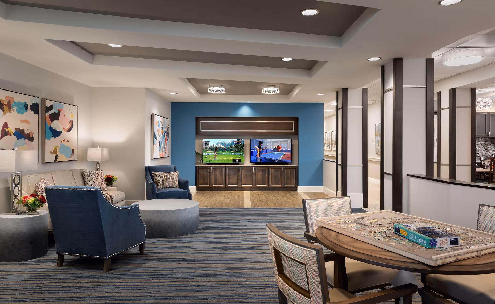 A spacious and modern living room area in an assisted living facility featuring comfortable seating including a beige sofa and blue armchairs, a round coffee table, and a wooden entertainment center with two flat-screen TVs mounted on a blue accent wall. The room is decorated with abstract art on the walls and has a puzzle table with chairs in the foreground. The space is well-lit with recessed ceiling lights and has a striped carpet floor.