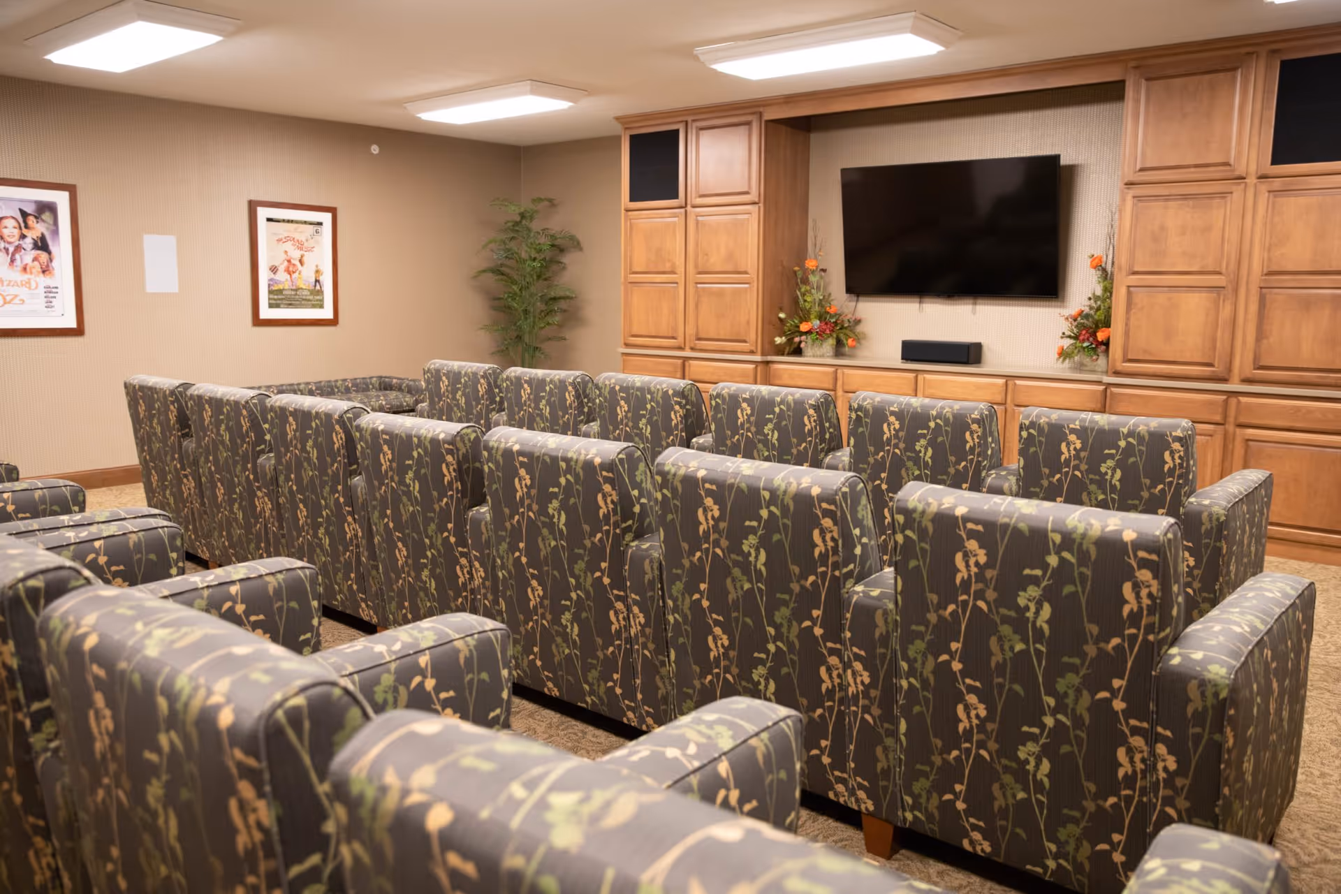 A cozy media room with multiple rows of patterned armchairs facing a wall-mounted flat screen TV. The room has wooden cabinetry on either side of the TV and framed movie posters on the walls. There are also two floral arrangements placed on the cabinet beneath the TV.