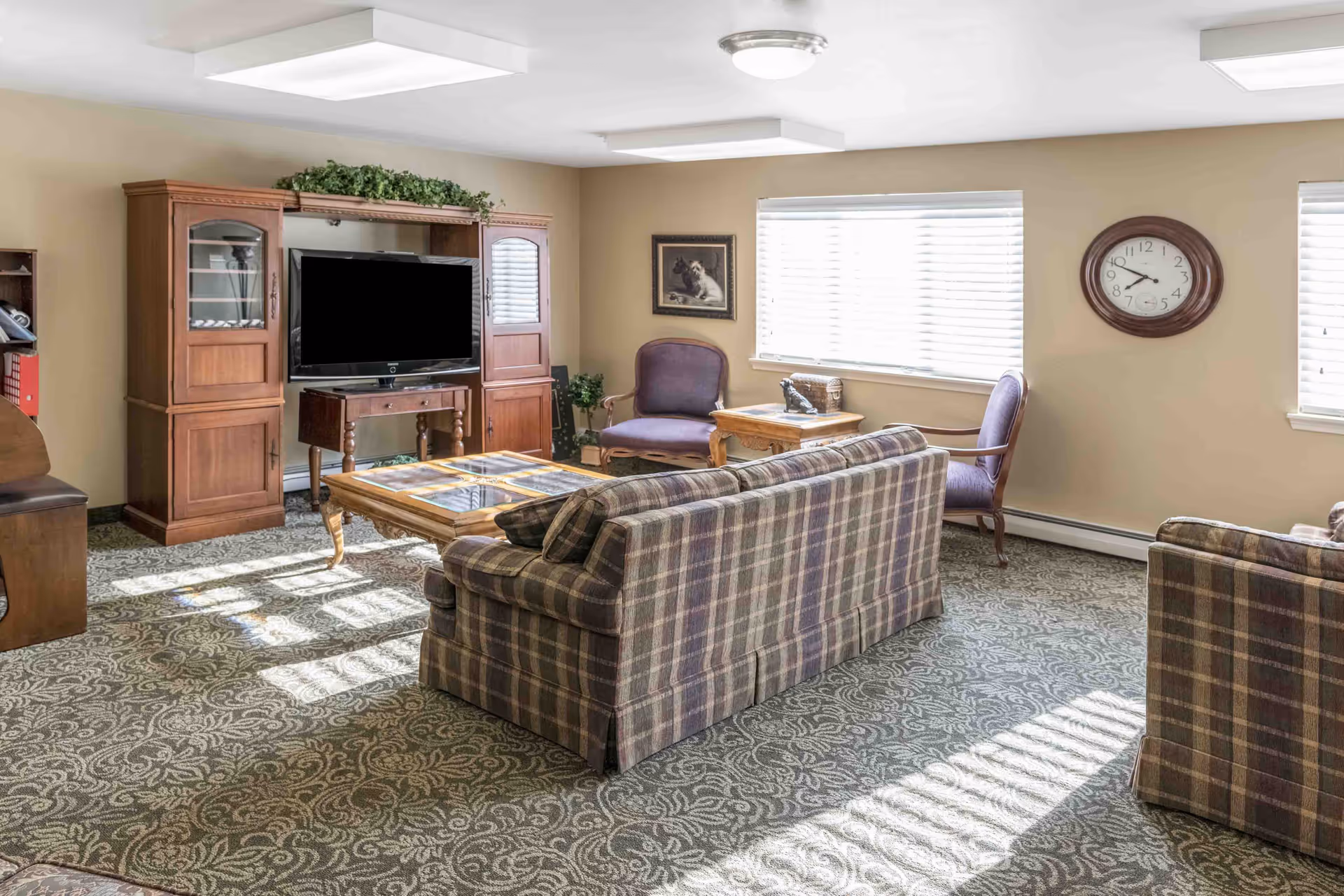 Sunlit common living room with plaid sofas, armchairs, a TV in a wooden cabinet, coffee table and wall clock.
