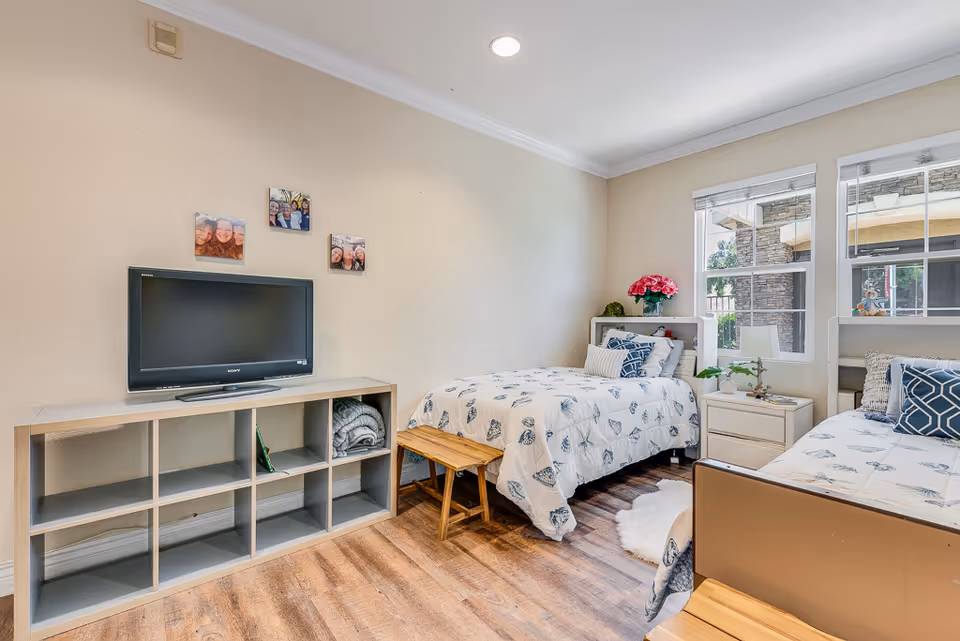 Bright shared bedroom with two twin beds, a TV on a shelving unit, bedside tables, and windows letting in natural light.