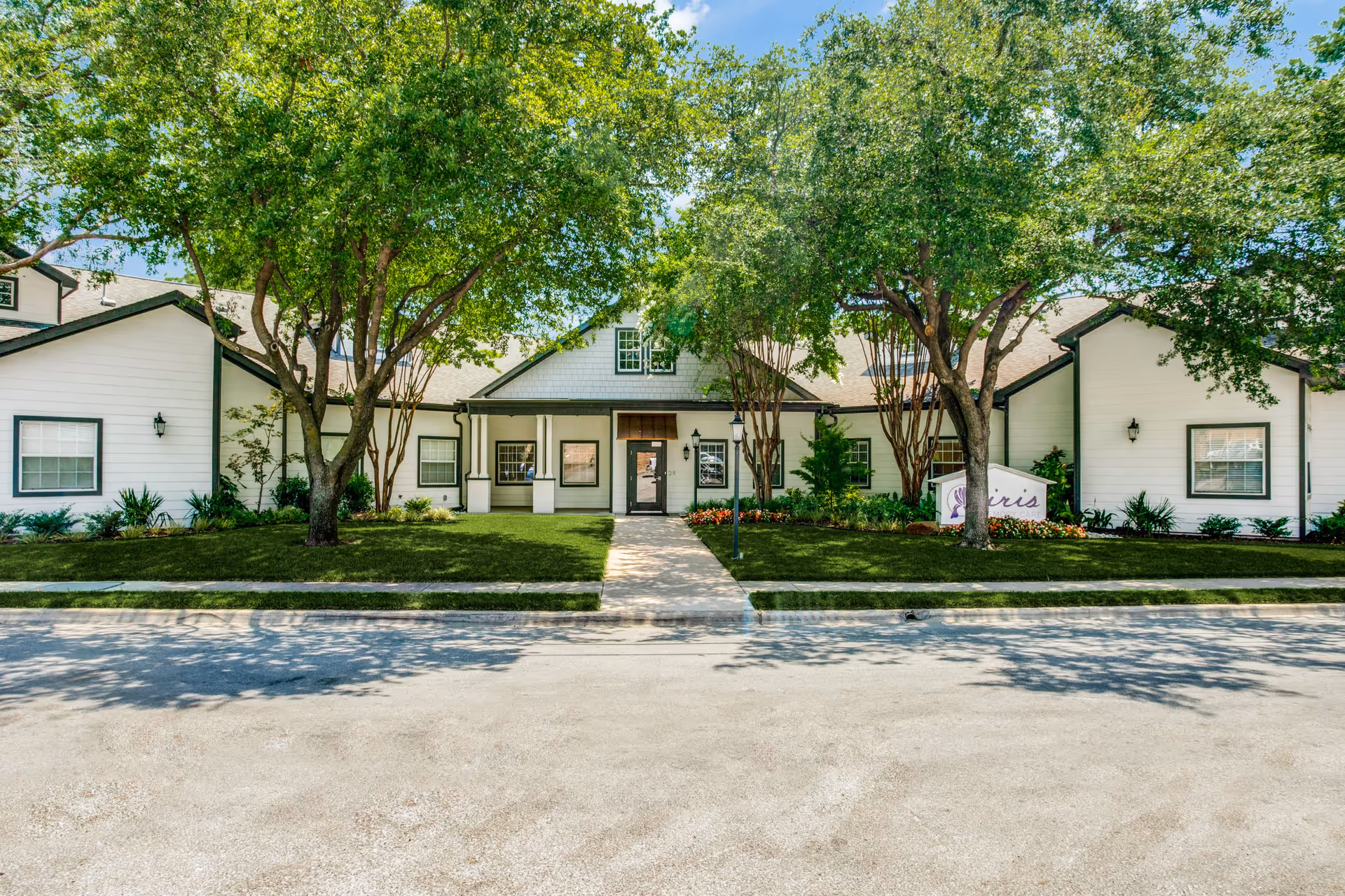 Front exterior view of a single-story building with white siding and dark trim, surrounded by green trees and landscaping, with a sidewalk leading to the entrance.
