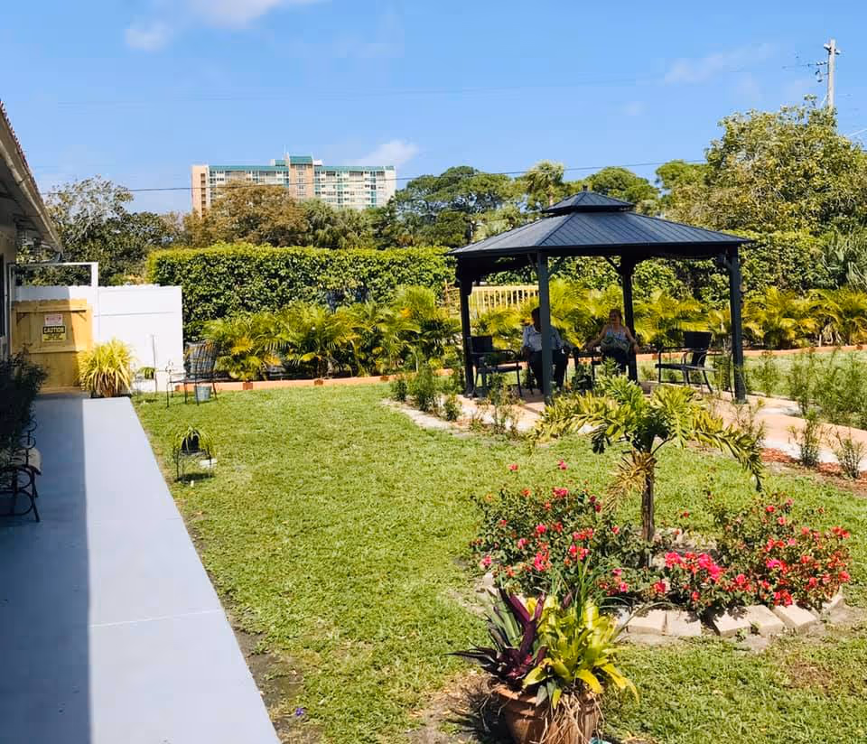 Outdoor garden area with green grass, a flower bed with red flowers, and a black gazebo where two people are sitting on chairs. There are bushes and trees in the background under a clear blue sky.