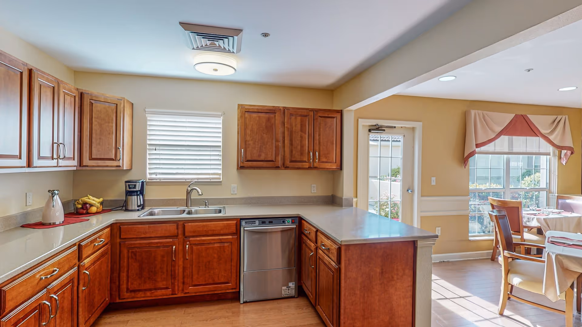 A bright kitchen area with wooden cabinets, a double sink under a window with blinds, a dishwasher, and a countertop extending into a dining area with tables and chairs near large windows with valances.