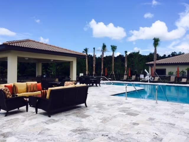Outdoor pool area with lounge chairs and seating under a covered patio. The pool is surrounded by a tiled deck with palm trees and a clear blue sky in the background.