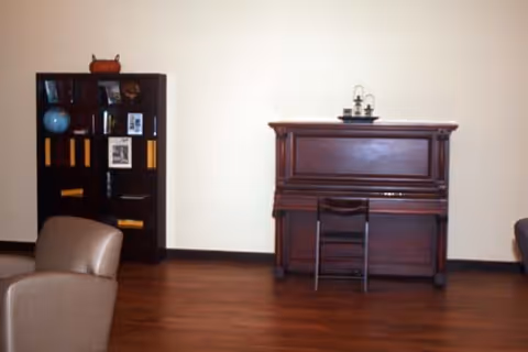 A room with wooden flooring featuring a dark wooden upright piano against a plain wall with a small chair in front of it. To the left, there is a dark wooden bookshelf with various items including a globe, framed pictures, and decorative objects. Part of a beige armchair is visible in the foreground.