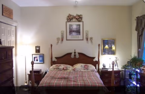 A cozy bedroom with a wooden four-poster bed covered with a plaid bedspread and floral pillows. There are two wooden nightstands on either side of the bed, each with a lamp and decorative items. A dresser is visible on the left side of the room, and framed pictures and artwork hang on the walls. A window with curtains is on the right side, letting in natural light.