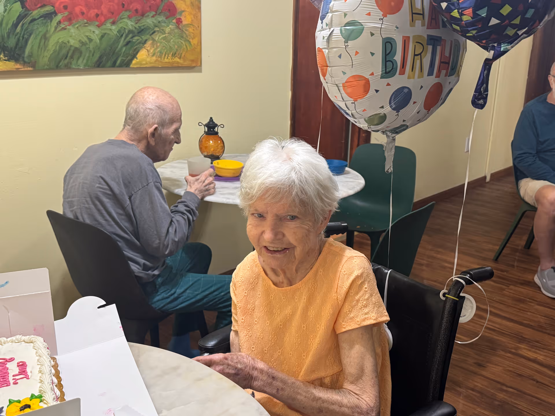 An elderly woman in an orange shirt sitting in a wheelchair at a table with a birthday cake and a 'Happy Birthday' balloon. In the background, an elderly man is seated at another table drinking from a cup. The room has wooden floors, light-colored walls, and a colorful painting on the wall.