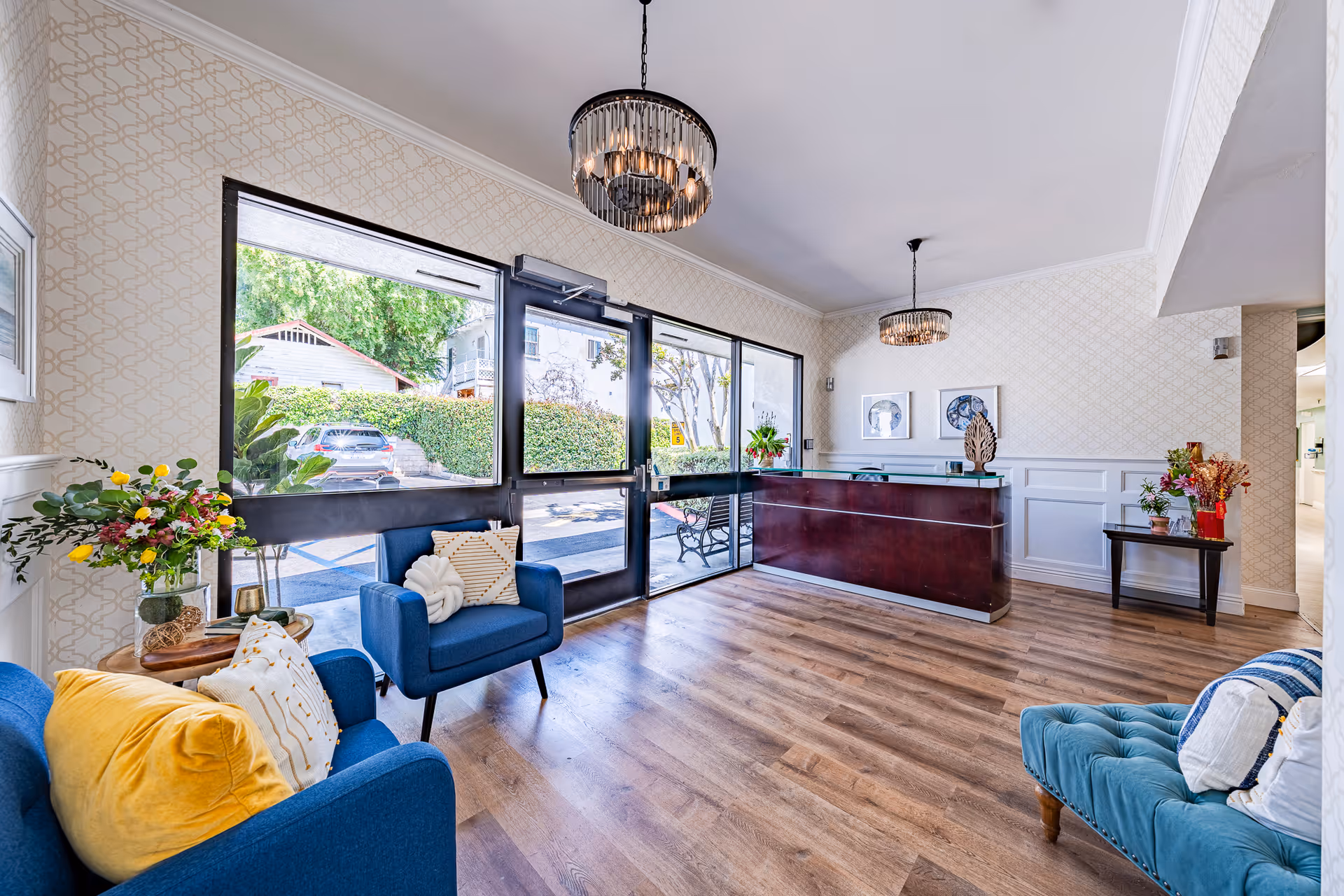A bright and welcoming reception area with large glass doors and windows letting in natural light. The room features a dark wooden reception desk, two blue armchairs with decorative pillows, a teal tufted bench, wooden flooring, and floral arrangements on side tables. Two modern chandeliers hang from the ceiling, and framed artwork is displayed on the walls.