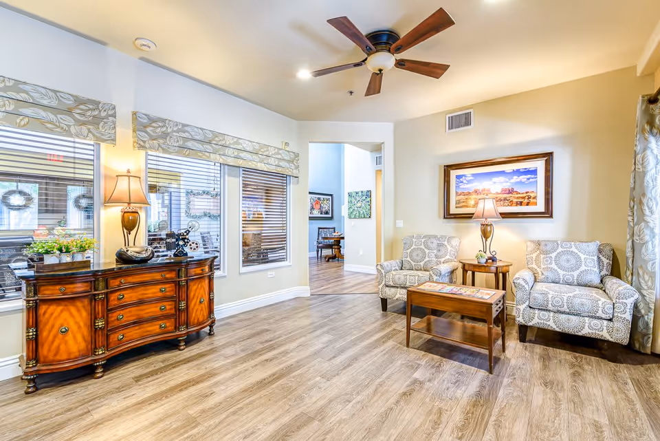 A bright and welcoming living room area in a senior living facility with two patterned armchairs, a wooden coffee table, and a side table with a lamp. There is a framed landscape picture on the wall, a ceiling fan with wooden blades, and a wooden sideboard with decorative items and a lamp near the windows with patterned valances.
