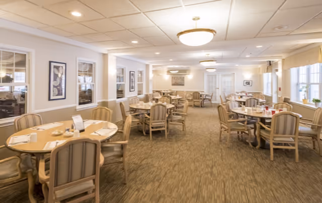 A spacious dining room with multiple round tables and cushioned chairs arranged neatly. The room is well-lit with ceiling lights and large windows on one side letting in natural light. The walls are decorated with framed artwork and the tables are set with placemats, cups, and condiments.