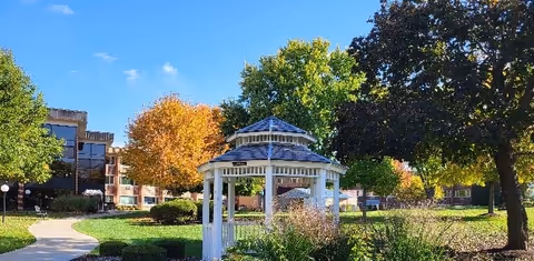 A bright outdoor garden area at Fairhaven Nursing Home featuring a white gazebo surrounded by green grass, bushes, and trees with autumn-colored leaves under a clear blue sky.