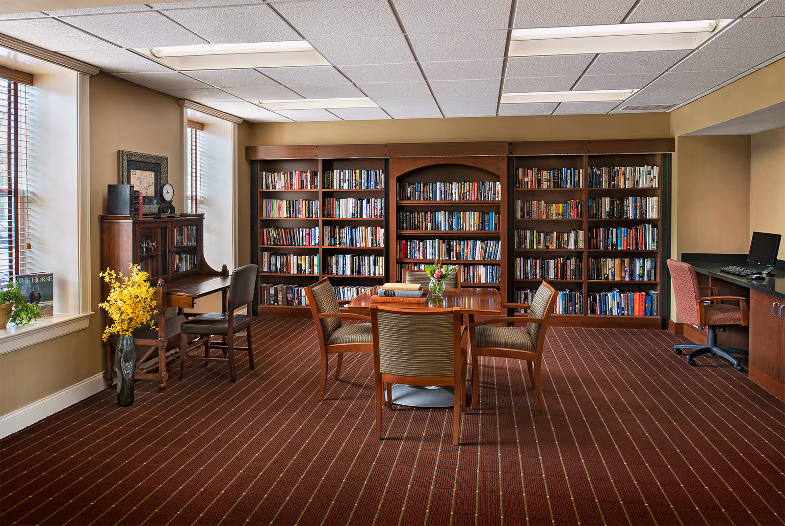 A cozy library room with large wooden bookshelves filled with books along the back wall. In the center, there is a wooden table with four chairs around it, and a small vase with flowers on the table. To the left, there is a wooden desk with a chair, a clock, and decorative items on top. Two large windows with blinds allow natural light into the room. On the right side, there is a computer workstation with a chair. The room has a brown carpet with a striped pattern and beige walls.